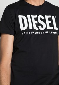 Černé tričko s krátkým rukávem s velkým bílým nápisem "DIESEL" a menším textem "FOR SUCCESSFUL LIVING", které má na sobě osoba.