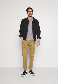 Chaqueta de mezclilla negra, camiseta gris, pantalones cargo color crema con bolsillos y zapatillas negras con detalles blancos. Líneas limpias, ajuste informal.