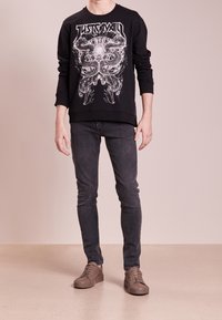 Pullover nero con design grafico, abbinato a jeans grigi scuri e sneakers marroni chiari. Il tessuto sembra morbido e ha una vestibilità comoda.