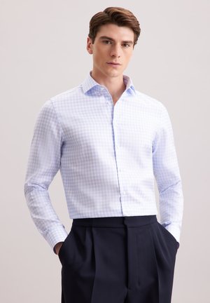 Camicia a scacchi blu chiaro e bianca con colletto a bottoni, maniche lunghe e tessuto morbido. Abbinata a pantaloni scuri, mani nelle tasche.