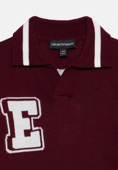 Polo en maille bordeaux avec des rayures blanches sur le col, grande lettre blanche E sur la poitrine et étiquette Emporio Armani à l’intérieur du col.