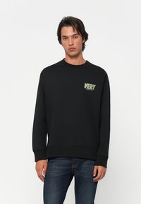 Νεαρός άνδρας φορώντας μαύρη φόρμα με το κείμενο "VERY" σε πράσινο και λευκό στο στήθος, συνδυασμένη με σκούρα μπλε τζιν, που στέκεται μπροστά σε λευκό φόντο.