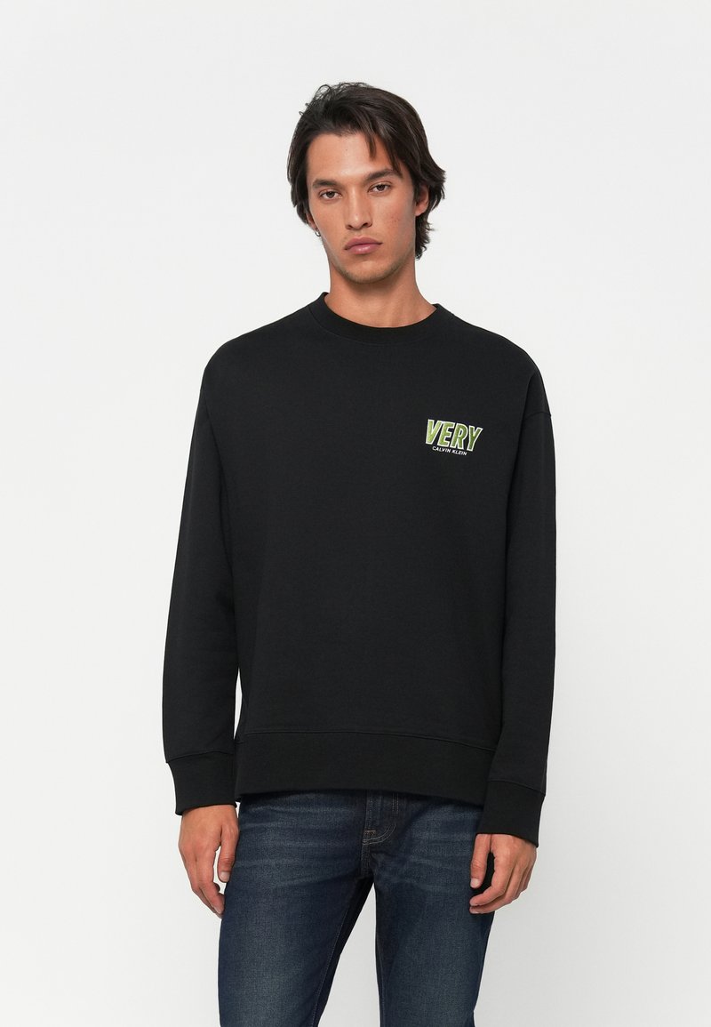 Νεαρός άνδρας φορώντας μαύρη φόρμα με το κείμενο "VERY" σε πράσινο και λευκό στο στήθος, συνδυασμένη με σκούρα μπλε τζιν, που στέκεται μπροστά σε λευκό φόντο.