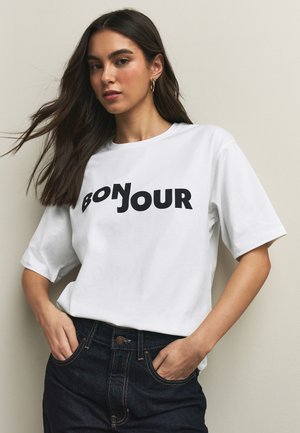 Mujer con pelo largo y castaño lleva una camiseta blanca con texto negro en negrita "BONJOUR" y vaqueros azul oscuro, de pie frente a un fondo beige.