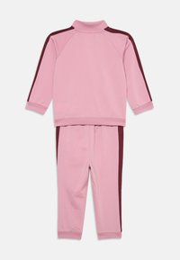 Roze sporttracksuit met hoge kraag, met bordeauxrode biezen aan zowel de jas als de broek. Gemaakt van zachte, gladde stof.