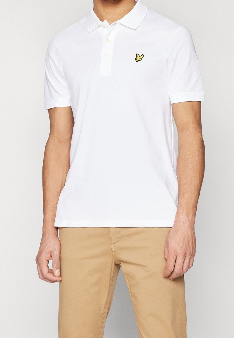 Polo blanc en coton avec une patte de boutonnage à trois boutons et un logo brodé jaune sur le côté gauche de la poitrine. Manches courtes, texture lisse.