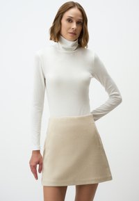Beige ausgestellter Rock aus weicher Stoffqualität mit glatter Textur und seitlicher Tasche, kombiniert mit einem figurbetonten weißen Langarm-Turtleneck-Oberteil.