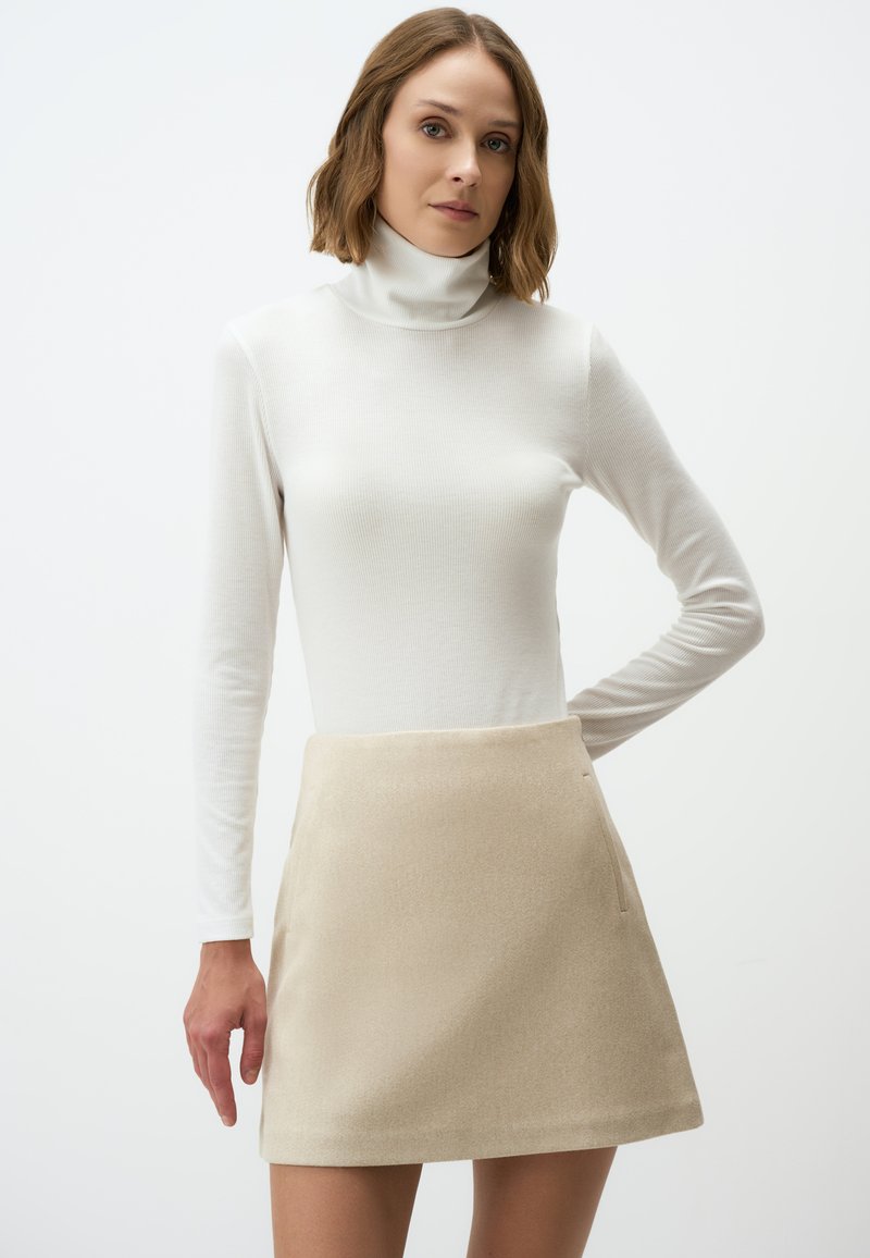 Beige ausgestellter Rock aus weicher Stoffqualität mit glatter Textur und seitlicher Tasche, kombiniert mit einem figurbetonten weißen Langarm-Turtleneck-Oberteil.