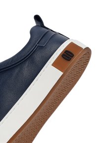 Granatowy skórzany sneaker z białą środkową podeszwą, brązową gumową podeszwą zewnętrzną z wzorem jodełki i czarnym logo na pasku z tyłu buta.