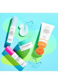 Four skincare products on a blue and green background: a blue spray, a pink spray, a white jar with a turquoise lid, and a white tube with an orange cap.