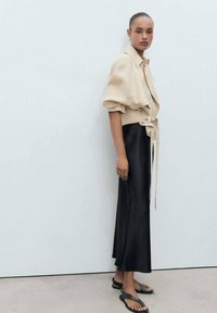 Chemise beige courte avec des manches bouffantes et une ceinture à la taille, associée à une jupe midi noire en satin et des sandales plates noires.