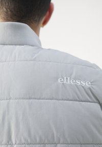 Svijetlosiva pufasta jakna s rebrastim ovratnikom i bijelim izvezenim "ellesse" logotipom na leđima, koja ima kviltanu teksturu i glatki materijal.