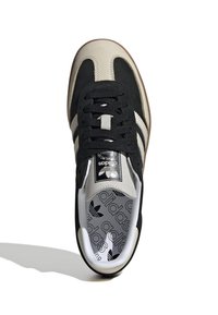 Svart och beige Adidas-sneaker med svarta snörningar, vita ränder på sidorna och Adidas-logotyp på tungan och innersulan, sedd ovanifrån.