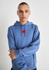 Felpa blu con un logo rettangolare rosso etichettato "HUGO". Presenta un cappuccio con cordini, polsini a coste e una vestibilità rilassata. Texture morbida.