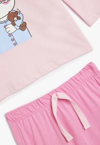 Juego de pijama de algodón de color rosa claro que presenta un gráfico de un personaje sosteniendo un oso de peluche marrón. Los pantalones tienen una suave cinturilla elástica con una cinta.