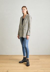 Blazer à carreaux beige et noir avec deux poches avant, associé à un jean bleu slim et des bottines noires, porté avec des lunettes claires.