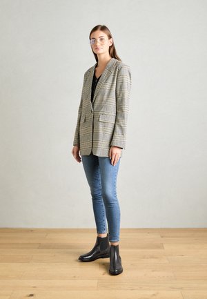 Blazer xadrez em bege e preto com dois bolsos frontais, combinado com jeans azul slim e botas de tornozelo pretas, usado com óculos transparentes.