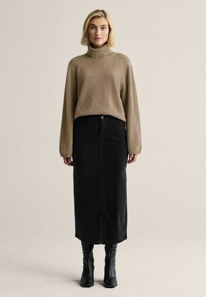 Nainen pukeutuneena vaaleanruskeaan pooloneuleeseen, mustaan midihameeseen ja mustiin saappaisiin seisomassa yksivärisen vaalean taustan edessä.