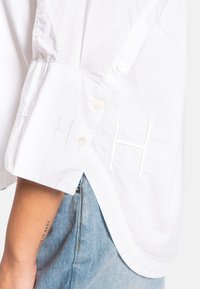 Hvit langermet skjorte med knappdetaljer på mansjettene, med broderte "H"-detaljer, laget av myk bomull.