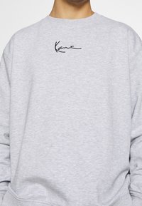 Grijze sweatstof trui van zachte stof, met een losse pasvorm, ronde hals en een zwart geborduurd handtekeninglogo op de borst.