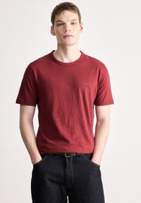 Κόκκινο βαμβακερό μπλουζάκι με κοντά μανίκια, στρογγυλή λαιμόκοψη, διακριτική σήμανση στο στήθος, συνδυασμένο με σκούρο τζιν και μαύρη ζώνη.