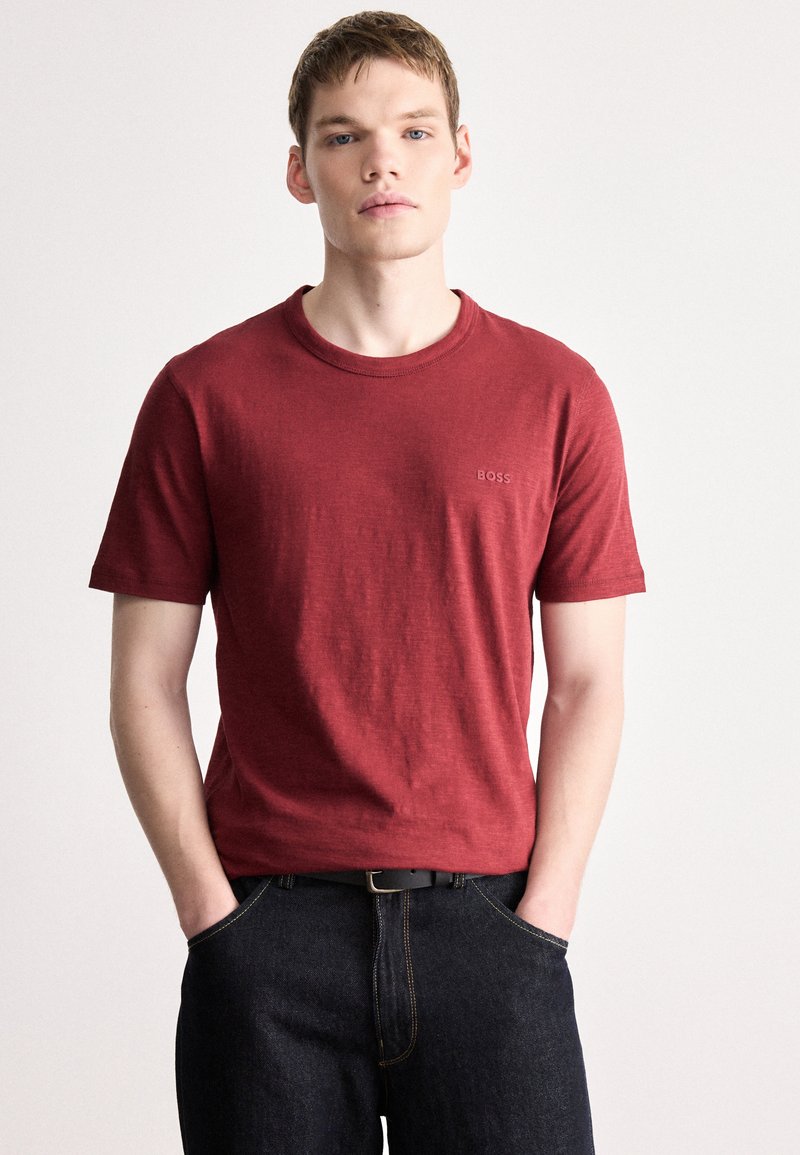 Κόκκινο βαμβακερό μπλουζάκι με κοντά μανίκια, στρογγυλή λαιμόκοψη, διακριτική σήμανση στο στήθος, συνδυασμένο με σκούρο τζιν και μαύρη ζώνη.