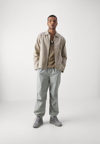Beige lightweight jacket, brown knitted sweater, loose pale green trousers, and gray sneakers. Features relaxed fit and minimalistic design.