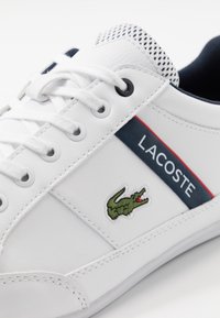 Baskets en cuir blanc avec des accents bleu marine et rouge, présentant un logo crocodile brodé vert et des lacets blancs, avec une languette en mesh texturé.