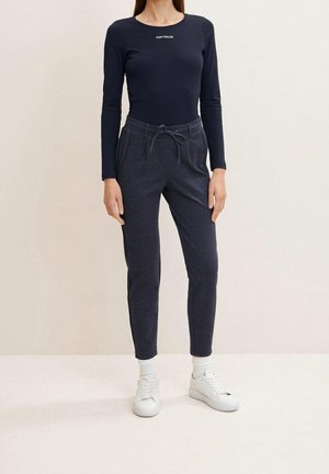 Kobieta ubrana w granatową, długorękawową koszulkę Tom Tailor, ciemnoszare spodnie ze ściągaczem, białe skarpetki i białe trampki, stojąca na tle jednolitego tła.