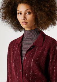 Μπορντό κα cardigan με υφή, που διαθέτει διακριτικές πούλιες και σχέδιο με γιακά. Φοριέται πάνω από μια ριγέ μπλούζα με ψηλό λαιμό.