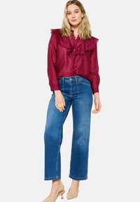 Maroon ruffled blouse, button-up front, paired with high-waisted blue denim jeans. Model wears beige heels, stands on a plain background.