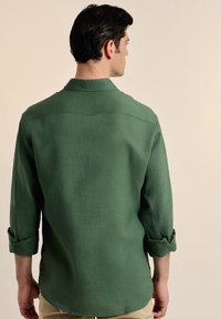 Camisa verde de manga larga hecha de un tejido texturizado. Presenta un cuello estándar y mangas arremangadas. El dobladillo es curvo y tiene un pequeño detalle de bucle.