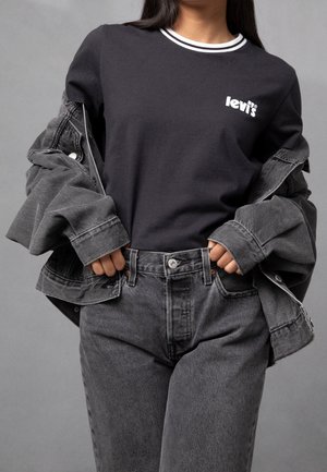 Młoda osoba w czarnej koszulce Levi’s, szarej dżinsowej kurtce zsuniętej z ramion i dopasowanych wysokich szarych dżinsach na szarym tle.