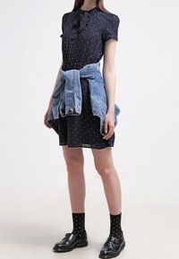 Mörkblå klänning med vita prickar, korta ärmar, åtsittande midja och knytkrage. Ljus denimjacka knuten runt midjan. Svarta skor och strumpor.