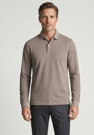 Man draagt een grijze poloshirt met lange mouwen en donkergrijze broek, staat met een neutrale uitdrukking voor een effen lichte achtergrond.