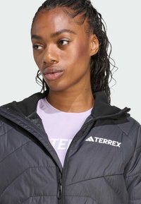 Mlada žena s pletenom kosom nosi crnu Terrex jaknu preko svijetlopurple košulje, promišljeno gleda u stranu na svijetloj pozadini.