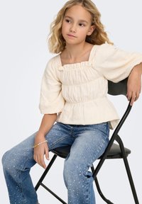 Šviesios grietinėlės spalvos tekstūruota palaidinė su surinktu kaklo iškirpimu ir pūstomis rankovėmis, derinama su išrankiomis mėlynomis džinsinėmis kelnėmis su sidabriniais blizgučiais, sėdinti ant juodos kėdės.