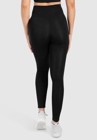 Leggings neri a vita alta con una trama liscia ed elasticizzata, caratterizzati da dettagli di cucitura lungo la parte posteriore e indossati con scarpe sportive bianche.