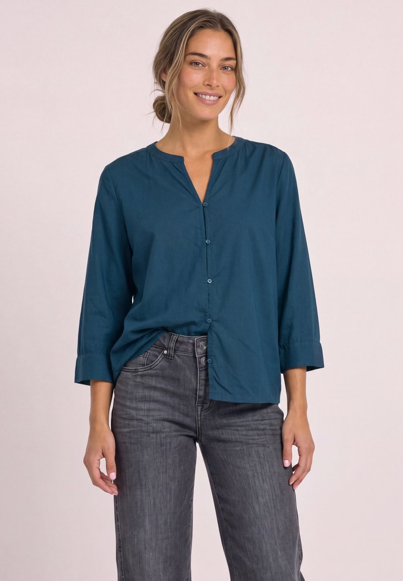 Femme souriante portant une chemise bleu sarcelle à boutons avec manches trois-quarts et un jean taille haute gris foncé devant un fond uni.