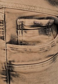 Jean en denim beige avec une poche arrière aux accents noirs décolorés. Le tissu a un aspect usé avec des détails de couture visibles.