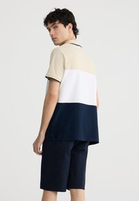 Polo shirt a blocchi di colore con sezioni beige, bianche e blu navy; maniche corte; colletto classico; abbinato a pantaloni corti blu navy con texture.