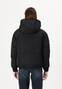 Svart dunjakke med hette, med quiltet design, elastiske mansjetter og innsnevret midje, brukt over blå jeans.