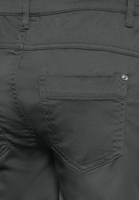 Pantaloni di tessuto grigio con una tasca posteriore di forma arrotondata, cuciture visibili e un accento di rivetto in metallo argentato. Texture liscia nel complesso.