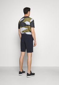 Conjunto deportivo para hombre que presenta una camiseta de manga corta con un patrón en tonos oliva y gris, combinada con pantalones cortos azul marino oscuros y zapatillas de deporte negras.