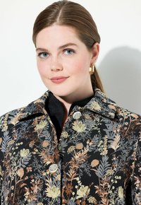 Mustat kukkakuosiset takki, jossa on kiiltävä pinta, monivärisiä kasvi- ja kukkasuunnitelmia, suuri kaulus ja kaksi pyöreää hopeapainiketta.