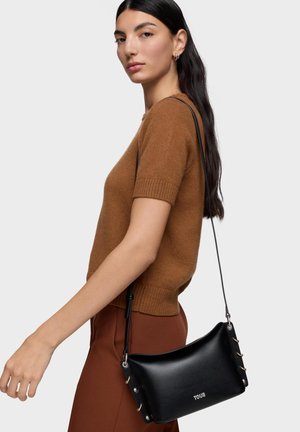 Mujer de cabello largo y oscuro, con un suéter marrón de manga corta y pantalones, llevando un bolso cruzado Tous de cuero negro sobre el hombro.