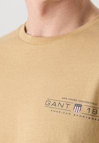 T-shirt à manches courtes, couleur beige, en coton doux avec un col rond. Présente un logo imprimé sombre et du texte sur le devant, mettant en avant les détails de la marque et de l'emplacement.