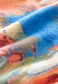 Farebný textil s mäkkou, chlpatou textúrou, ktorý obsahuje živé vzory v modrých, oranžových a zelených odtieňoch, vytvárajúci zmesitý, abstrahovaný dizajn.