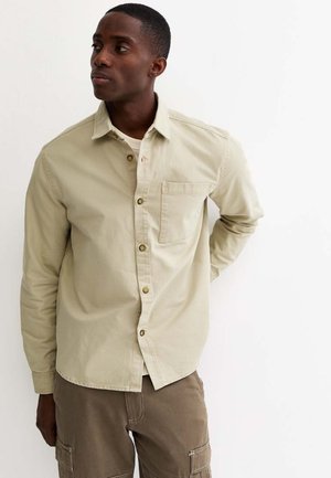 Młody mężczyzna o krótkich włosach, ubrany w beżową koszulę zapinaną na guziki, na białą koszulkę oraz brązowe spodnie typu cargo, stojący na tle białego tła.