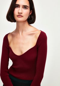 Top lungo a maniche lunghe color bordeaux con scollatura a V profonda, corpino aderente e tessuto testurizzato. Il modello è presentato in un contesto con sfondo neutro.